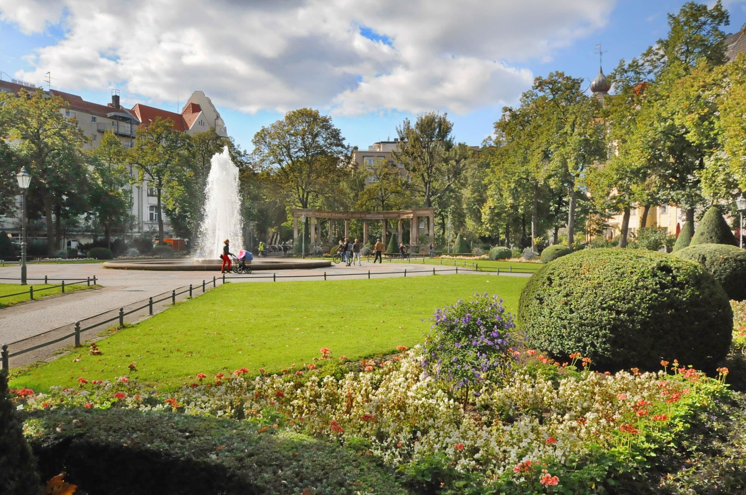 Parkanlage mit zentralem Springbrunnen, Rasenflächen, Blumenbeeten und Blick auf Bäume und Stadthäuser
