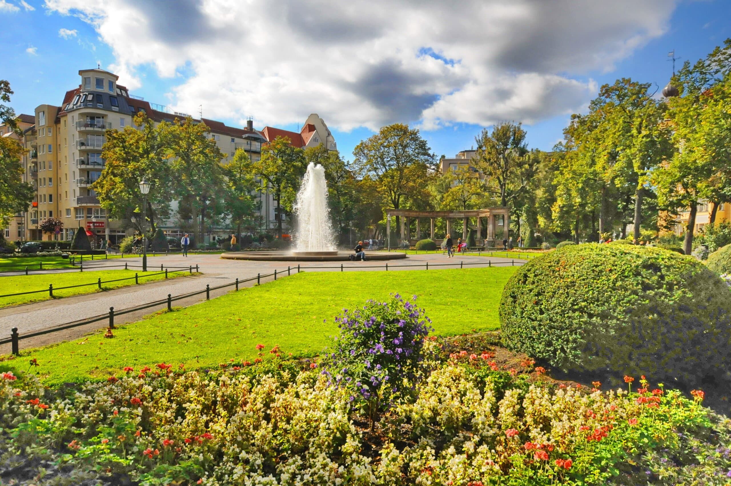 Blick über Blumenbeet auf städtischen Park mit Springbrunnen und historischen Wohnhäusern im Hintergrund