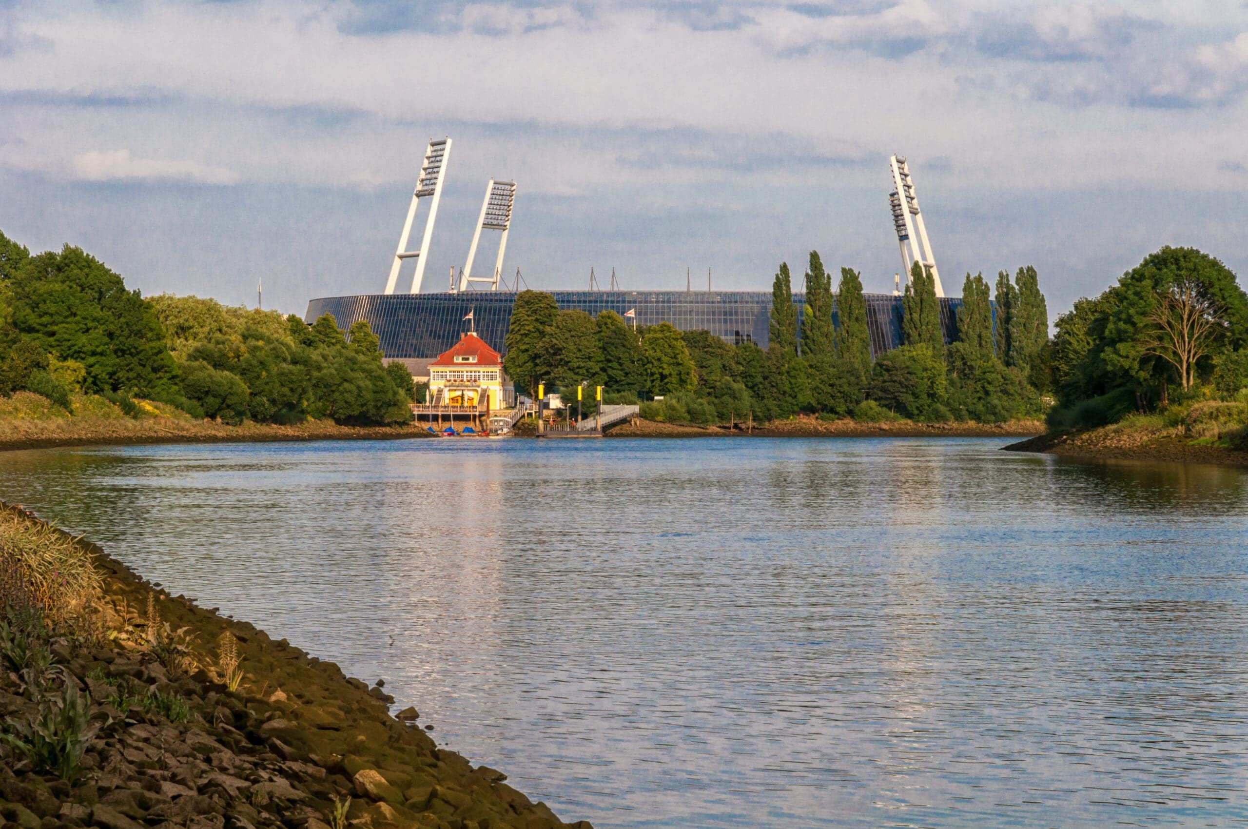 Sportstadion mit markanten Flutlichtmasten hinter Flussufer und Bootshaus aus der Uferperspektive