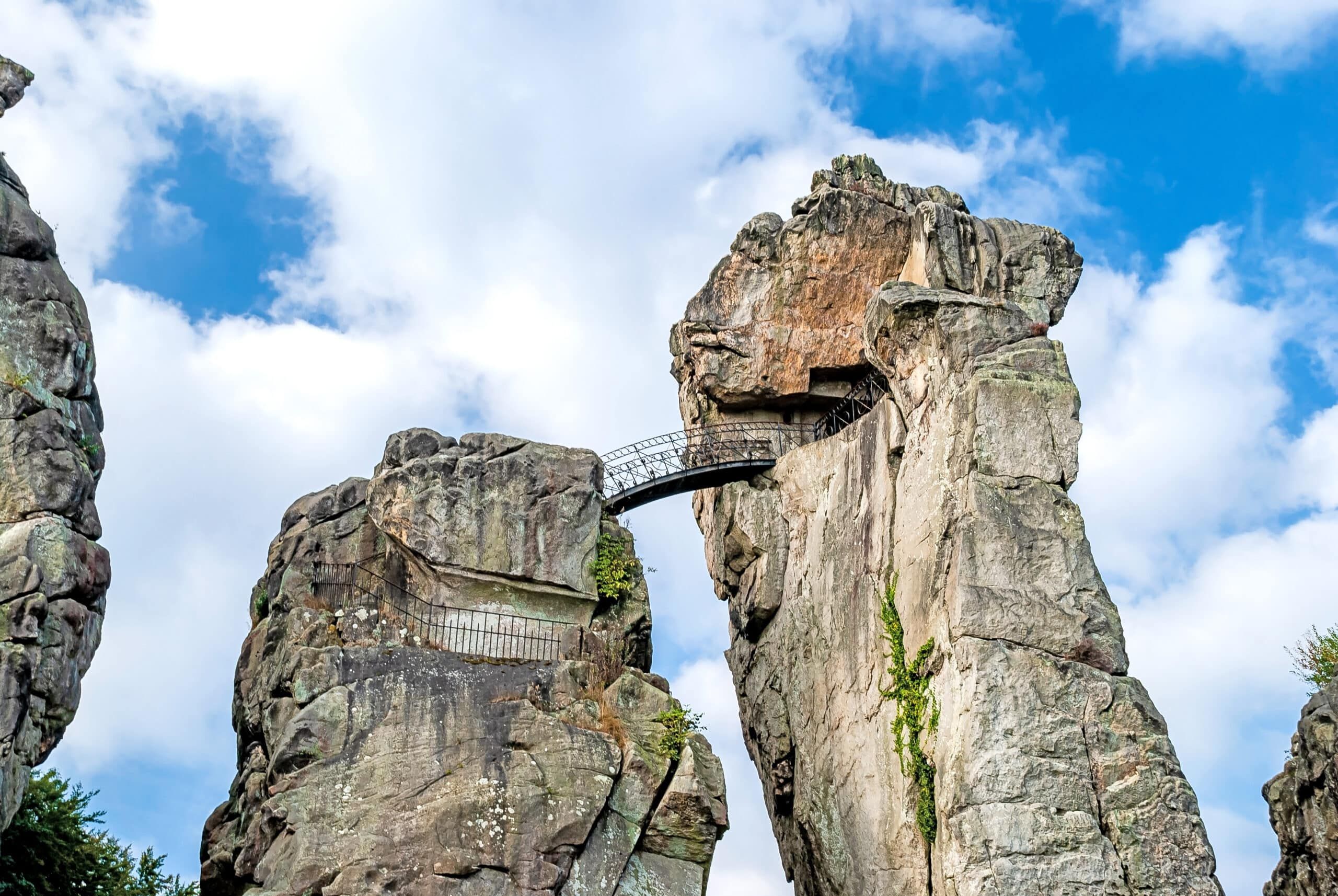 Seitliche Ansicht einer massiven Felsformation mit schmaler Stahlbrücke und teils begrünter Oberfläche