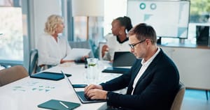 Bürobesprechung mit Geschäftspersonen am Tisch und Laptop vor großem Monitor mit Diagrammen