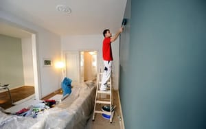 Maler auf Leiter streicht Wohnzimmerwand modern in Blaugrau mit abgedecktem Sofa und Werkzeug im hellen Altbauzimmer